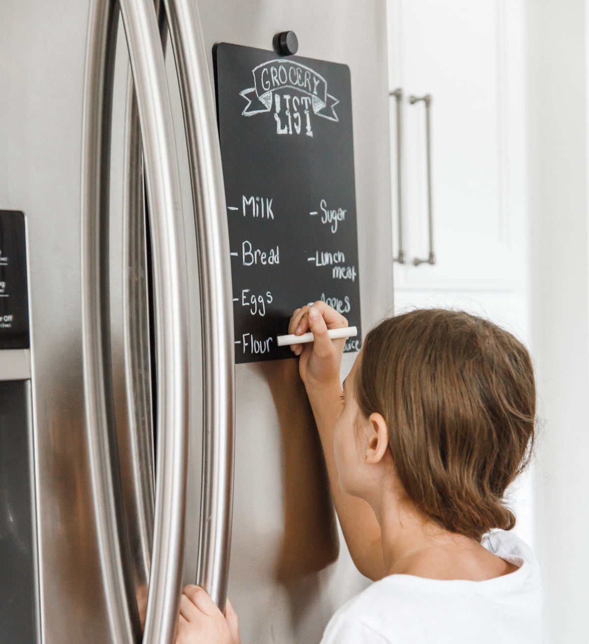 Child writing on a chalkboard attached to a refrigerator with a 'Grocery List' written on it.