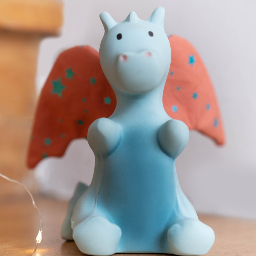 A small blue dragon figurine with orange wings decorated with blue stars, sitting on a wooden surface with warm fairy lights in the foreground.