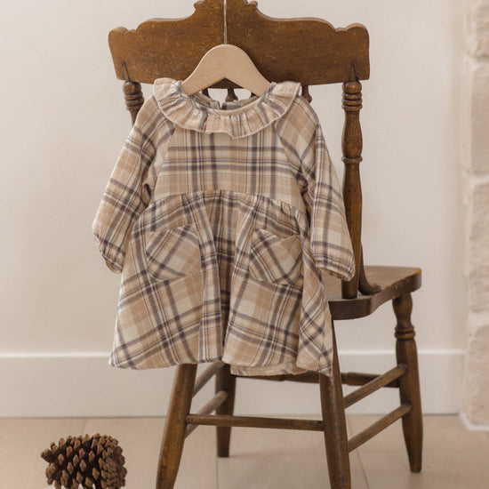 A toddler girl's long-sleeve plaid dress in beige, brown, and white hangs on a wooden hanger from a rustic wooden chair, with a pinecone and books on the floor nearby.