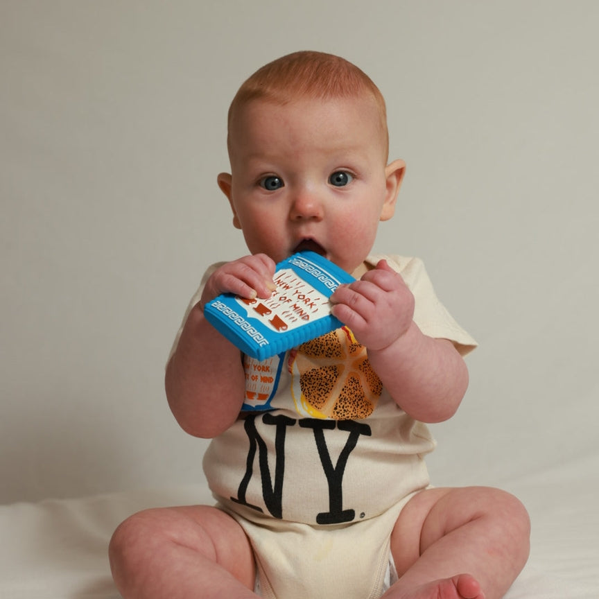 A baby sitting on a white surface against a light beige background, wearing a cream-colored onesie with 'NY' printed in black and holding a blue teething toy.