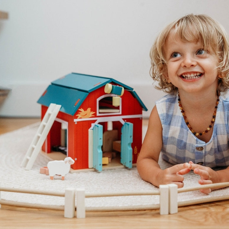 A young blonde-haired child, wearing a blue and white gingham sleeveless collared shirt, smiles happily while playing on the floor with the Wooden Big Red Barn Playset by Mentari, surrounded by the barn, fences, and small wooden animal figures.