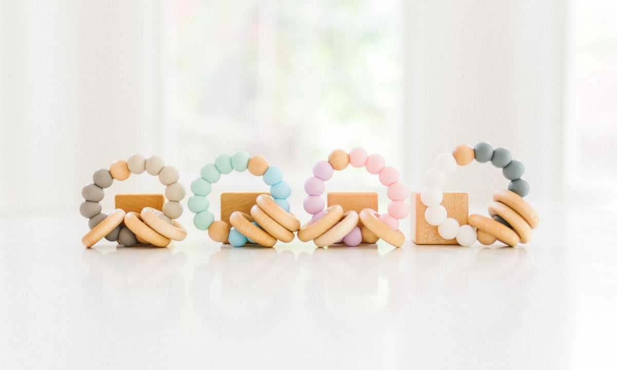Four pastel-colored wooden bead teething rings with wooden blocks, arranged in a row on a white reflective surface with a soft, blurred window background.