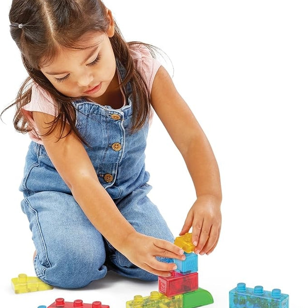 A young girl wearing denim overalls kneeling on a white background and stacking colorful, translucent Jelly Blox sensory blocks.