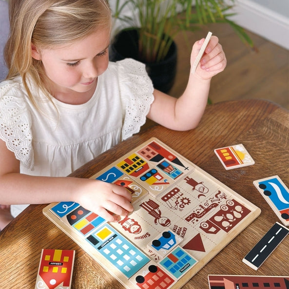A young girl with blonde hair tied back sits at a wooden table, smiling as she plays with the Mentari Wooden Town Puzzle, holding a puzzle piece in her hand while assembling the colorful town scene.