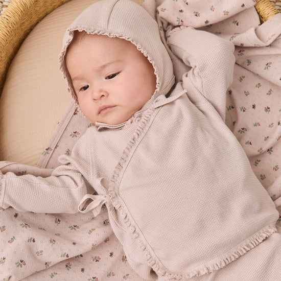 A baby wearing a soft blush pink hooded romper and matching floral blanket, lying on a woven mat.