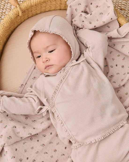 A baby wearing a soft blush pink hooded romper and matching floral blanket, lying on a woven mat.