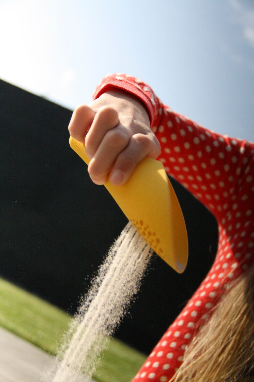 Person wearing a red polka dot dress holding a yellow sand toy with sand being poured out against a blurred background.