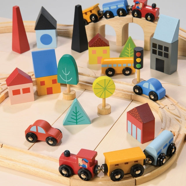 A top-down, angled view of a wooden toy train set on a light surface, featuring a curved wooden track layout with a blue train engine and yellow cargo cars. Colorful wooden town buildings, including a red house, blue structures, and abstract trees, surround the tracks alongside a yellow traffic light, a red toy car, and a yellow toy truck.
