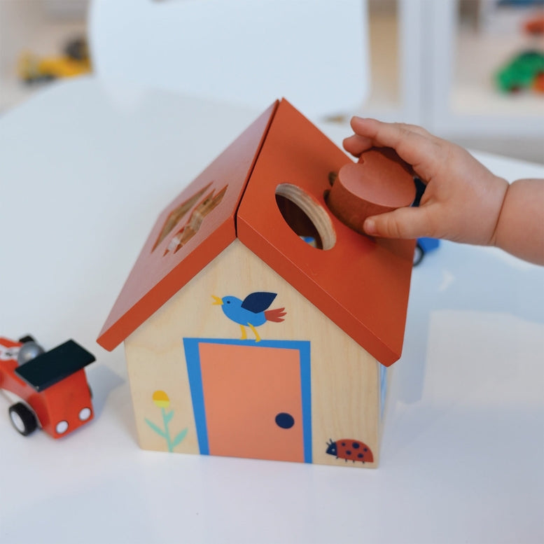A toddler's hand drops a wooden cylinder into the matching slot on the roof of a wooden house shape sorter.