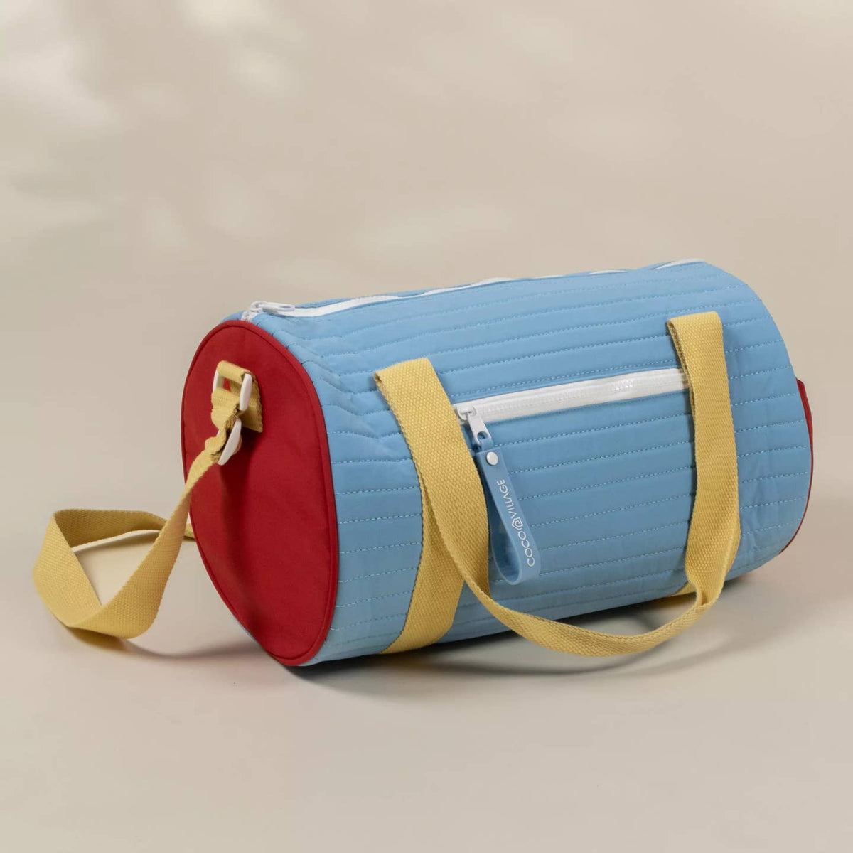 A light blue cylindrical duffel bag with red end caps, yellow straps, and a white zipper pocket, shown on a neutral beige background.
