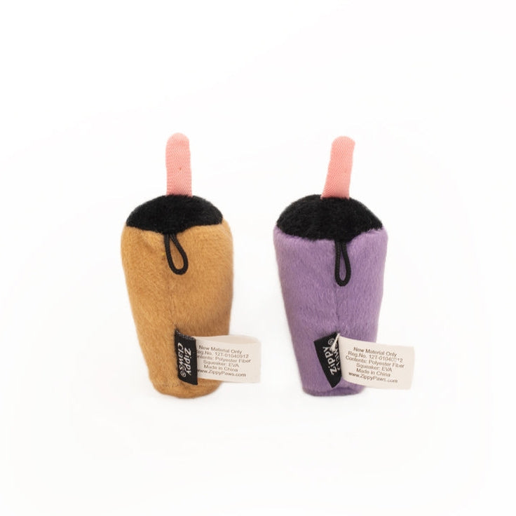 Two small plush drink cup toys on a plain white background. One is tan with a pink straw and black top, and the other is lavender-purple with a pink straw and black top.