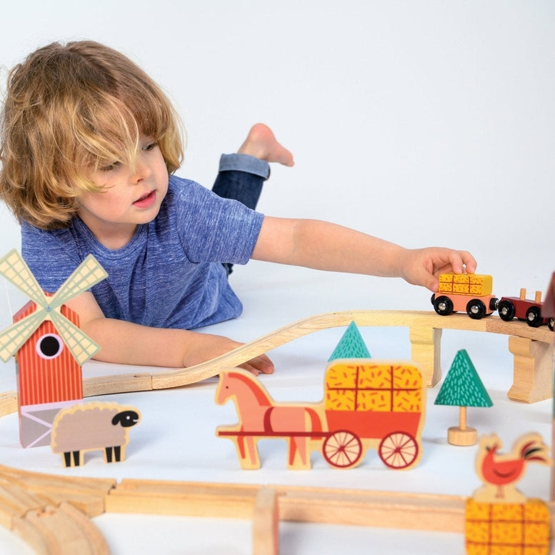A young child playing on the floor with a wooden farmyard-themed train set, placing a small wooden train onto the tracks next to a red barn and windmill.