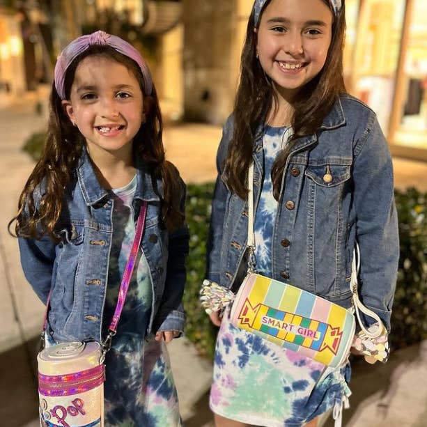 Two young girls wearing denim jackets and tie-dye outfits with colorful bags.