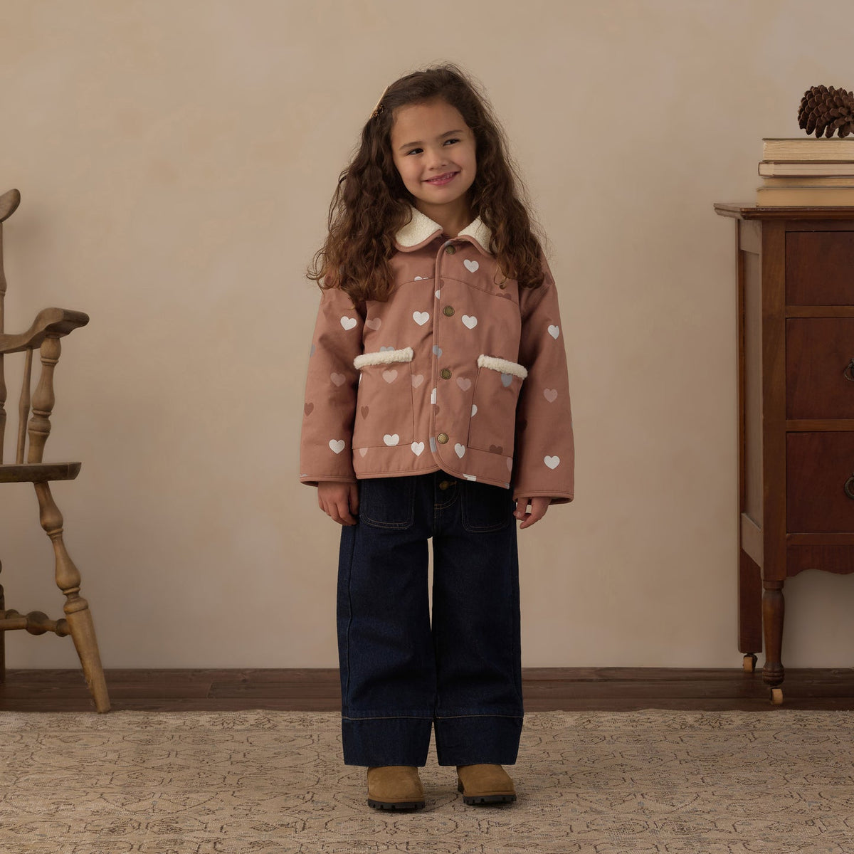 A young girl stands in a cozy indoor room wearing, a pink patterned sweater, dark blue wide-leg jeans, and pink shoes.