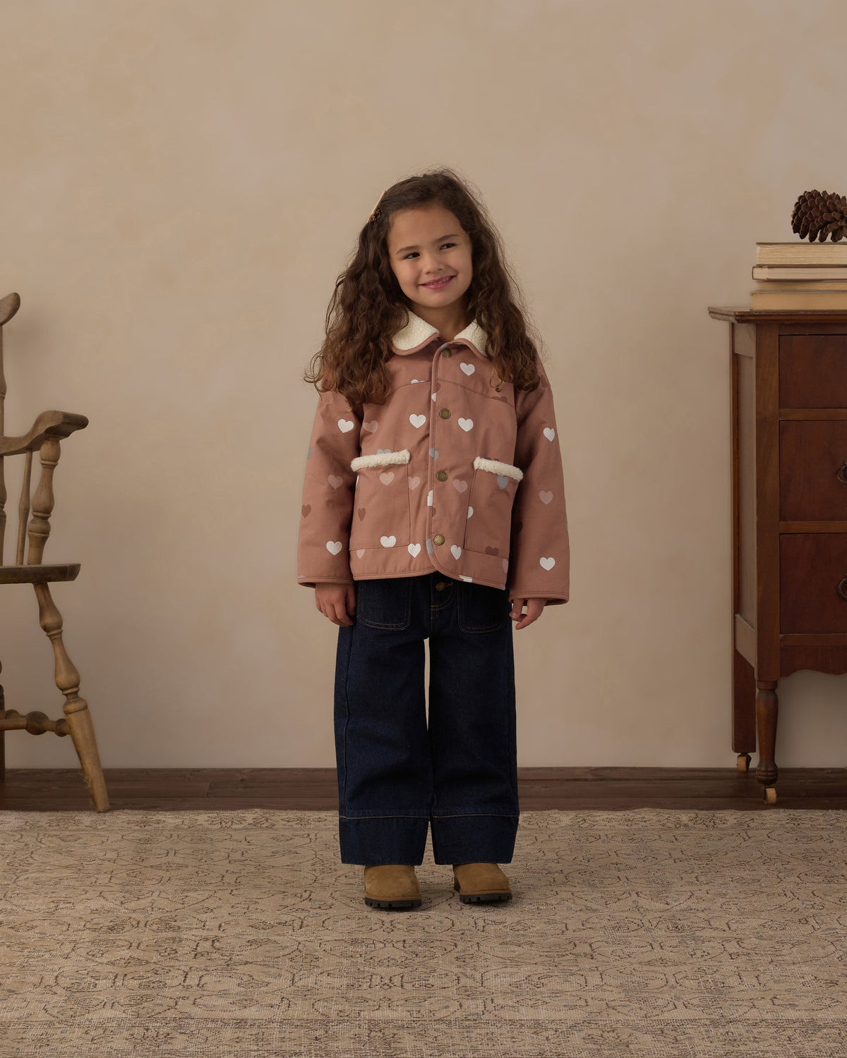 A young girl stands in a cozy indoor room wearing, a pink patterned sweater, dark blue wide-leg jeans, and pink shoes.