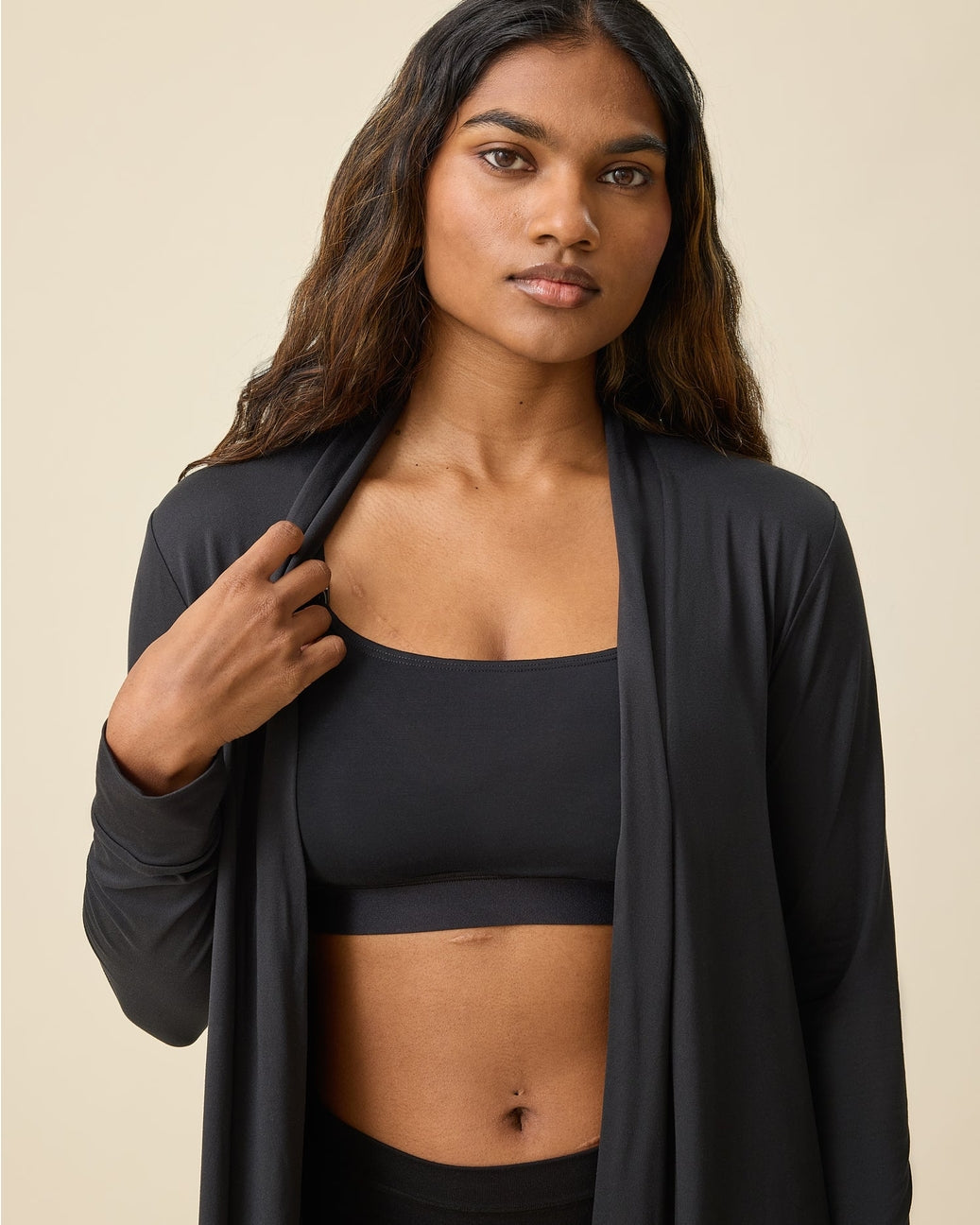 Woman wearing a black sports bra and leggings with a black cardigan on a beige background