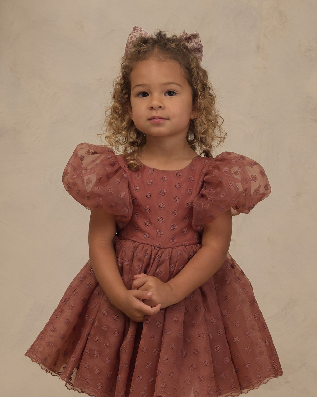 A young girl with curly light brown hair wearing a dusty rose pink tulle dress with puff sleeves and a matching bow headband, standing against a textured beige studio backdrop.