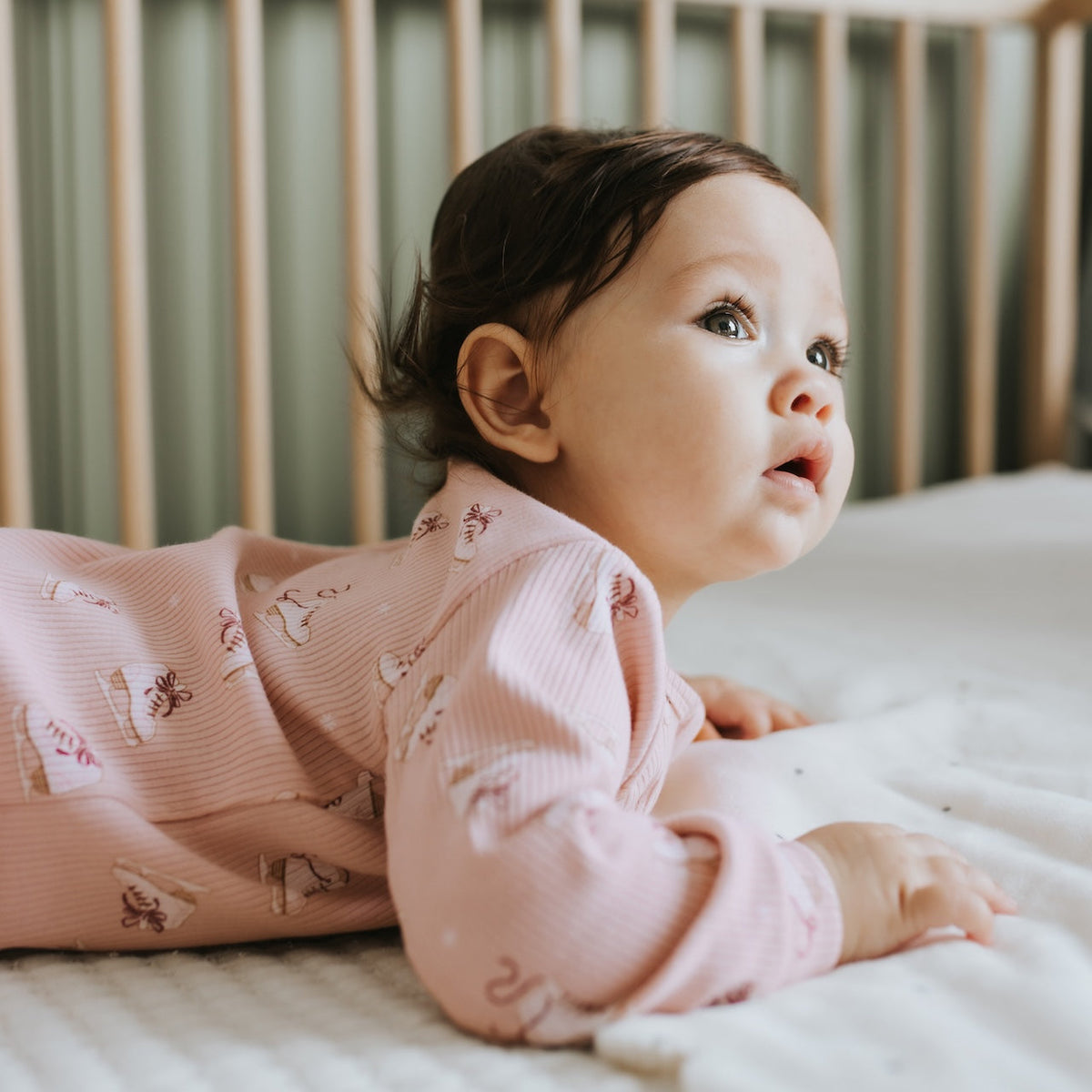 A baby wearing a pink ribbed long-sleeve romper with a small bunny print, lying on their stomach on a white quilted mattress in a crib.
