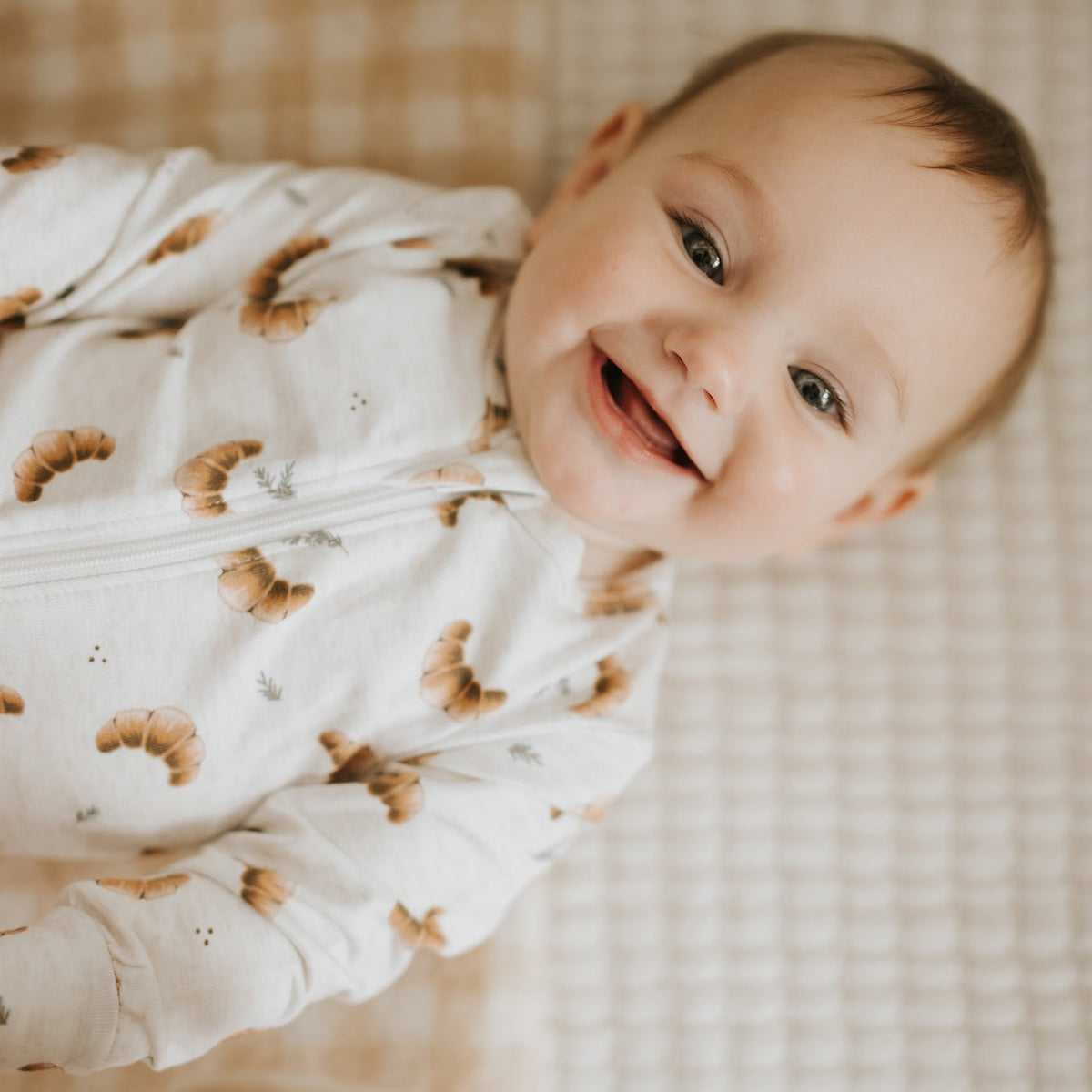 A smiling baby lying on a checkered blanket, wearing a white long-sleeve sleep sack with a brown crescent moon pattern.