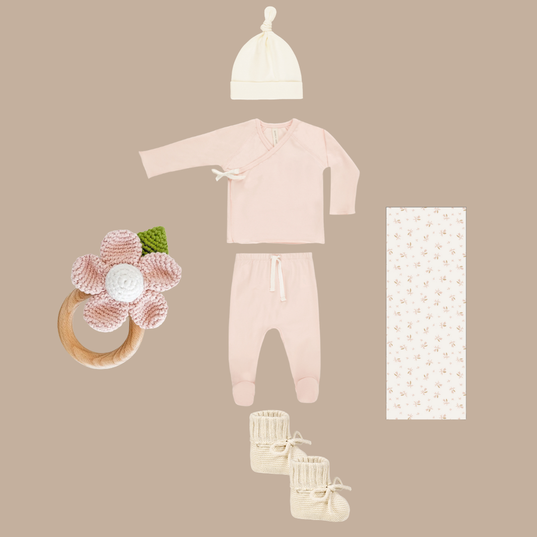 Baby clothing and accessories including a onesie, hat, booties, and a floral ring on a beige background.