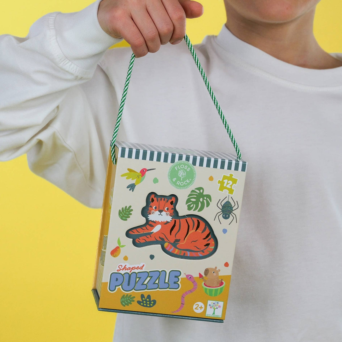 A person in a white sweatshirt holds a small illustrated puzzle box by a green-and-white rope handle against a bright yellow background. The box features a cartoon tiger, bird, spider, and the words 'Shipped PUZZLE'.