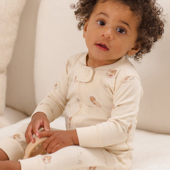 A baby with curly dark hair sits on a white sofa wearing a cream-colored long-sleeve footless sleeper with a zipper and a small brown animal print.