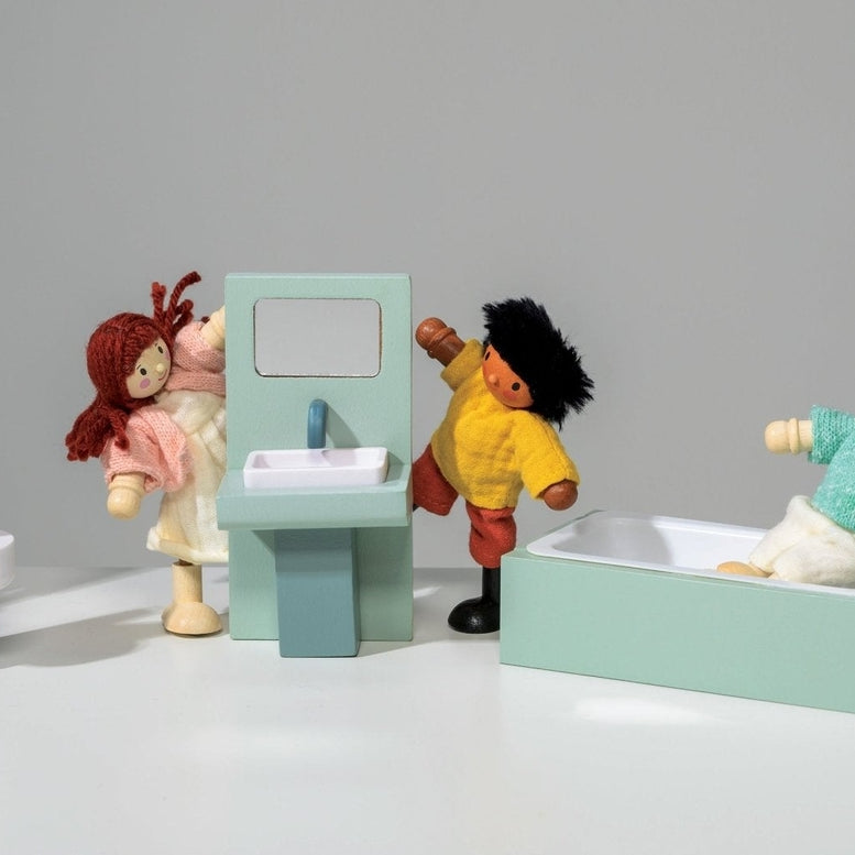 A lifestyle product photo featuring the Wooden Bathroom Furniture set by Mentari alongside several dollhouse dolls interacting with the pieces against a white background. One doll carries a wooden towel stand, another stands next to the toilet, a third washes their hands at the sink unit with a mirror, a fourth relaxes in the mint-green bathtub, and a fifth carries a pink fabric laundry basket.