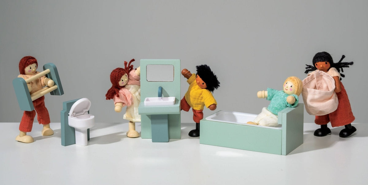 A lifestyle product photo featuring the Wooden Bathroom Furniture set by Mentari alongside several dollhouse dolls interacting with the pieces against a white background. One doll carries a wooden towel stand, another stands next to the toilet, a third washes their hands at the sink unit with a mirror, a fourth relaxes in the mint-green bathtub, and a fifth carries a pink fabric laundry basket.