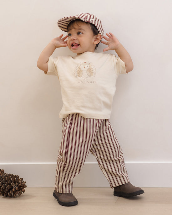A toddler stands against a plain light wall wearing a cream short-sleeve T-shirt with a turkey graphic and the words 'LITTLE TURTLE', striped pants in cream and dark red, a matching striped cap, and dark brown shoes.