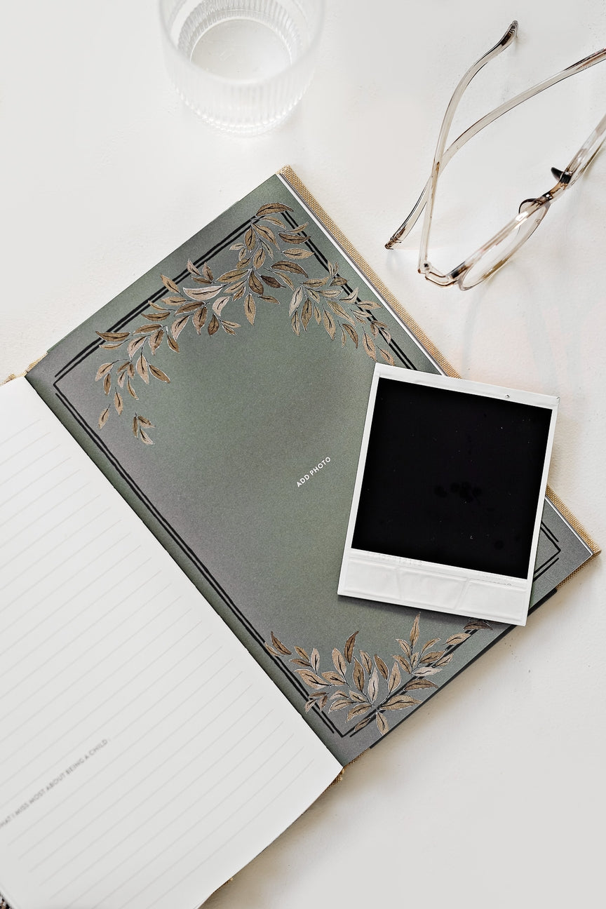 Green notebook with floral design on cover, white photo frame, and glasses on a light surface