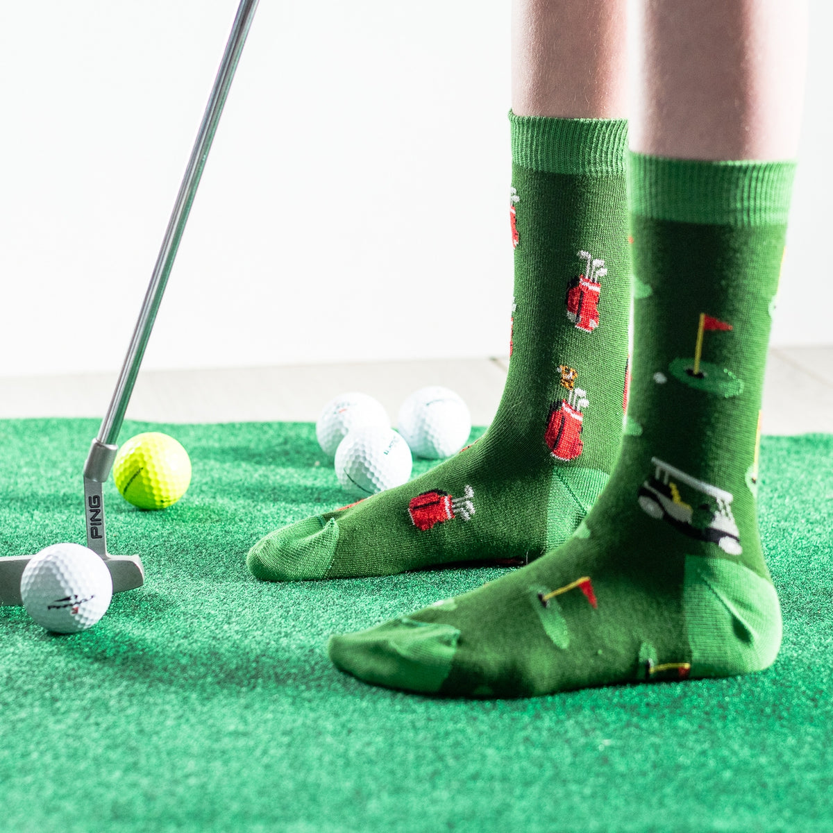 A person wearing green novelty golf socks with illustrated golfers and clubs, standing on a green turf mat with golf balls and a putter nearby.