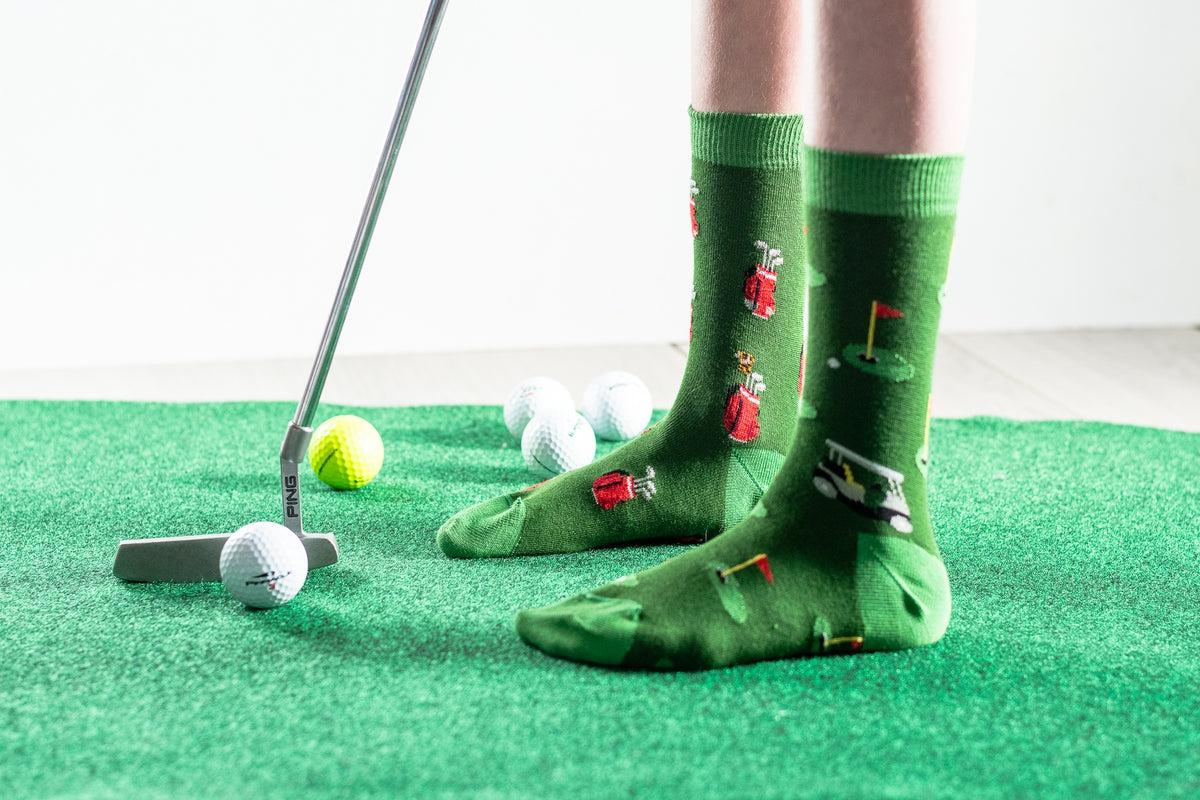 A person wearing green novelty golf socks with illustrated golfers and clubs, standing on a green turf mat with golf balls and a putter nearby.