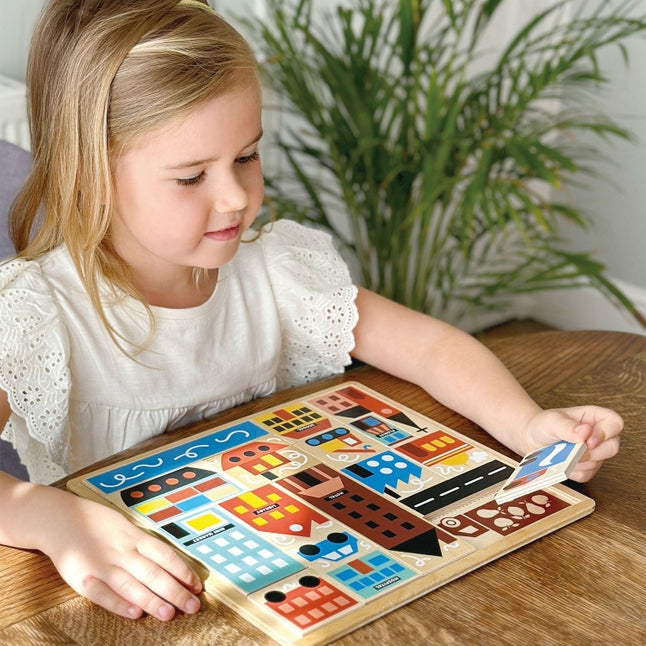A young girl with blonde hair tied back sits at a wooden table, smiling as she plays with the Mentari Wooden Town Puzzle, holding a puzzle piece in her hand while assembling the colorful town scene.