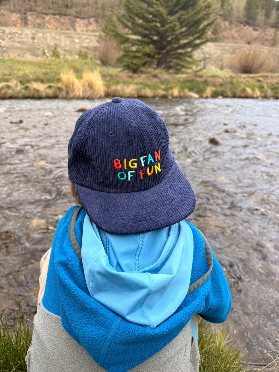 Person wearing a blue cap with colorful text, standing by a river.