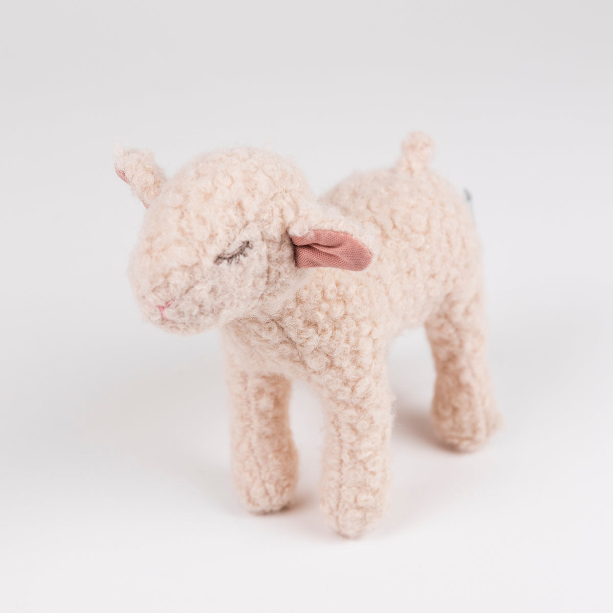 A small plush sheep toy with curly cream-colored wool, closed embroidered eyes, pink inner ears, and a pink nose, standing on a clean white background.