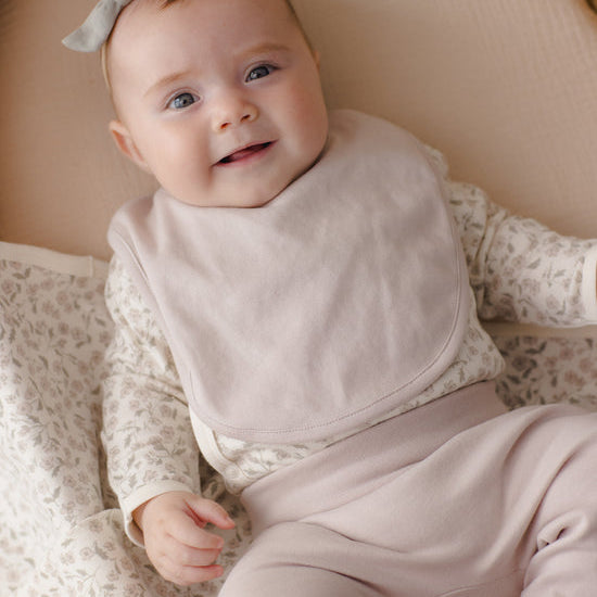 A smiling baby wearing a light beige bib over a matching beige and white patterned long-sleeve top and beige pants, lying on a soft beige blanket.