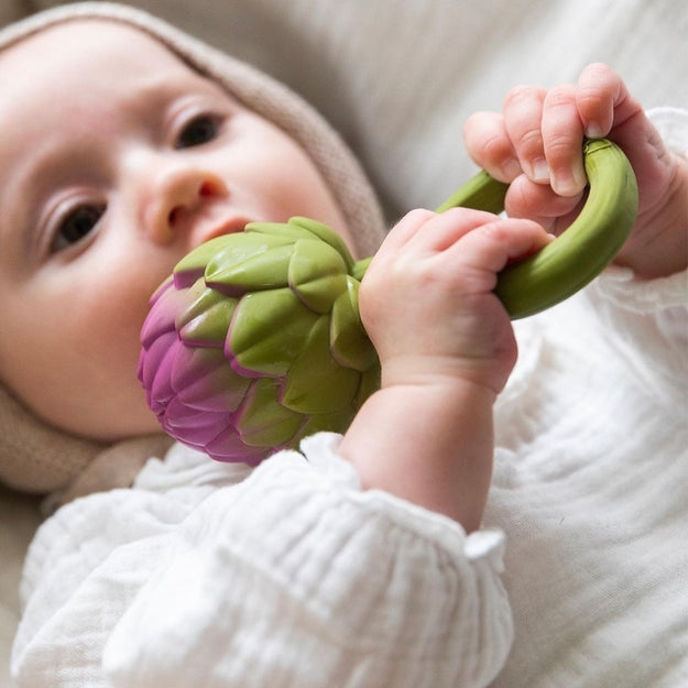 Baby lying down in a white ruffled outfit, holding the pink-and-green Oli & Carol artichoke rattle toy and exploring it with their hands.