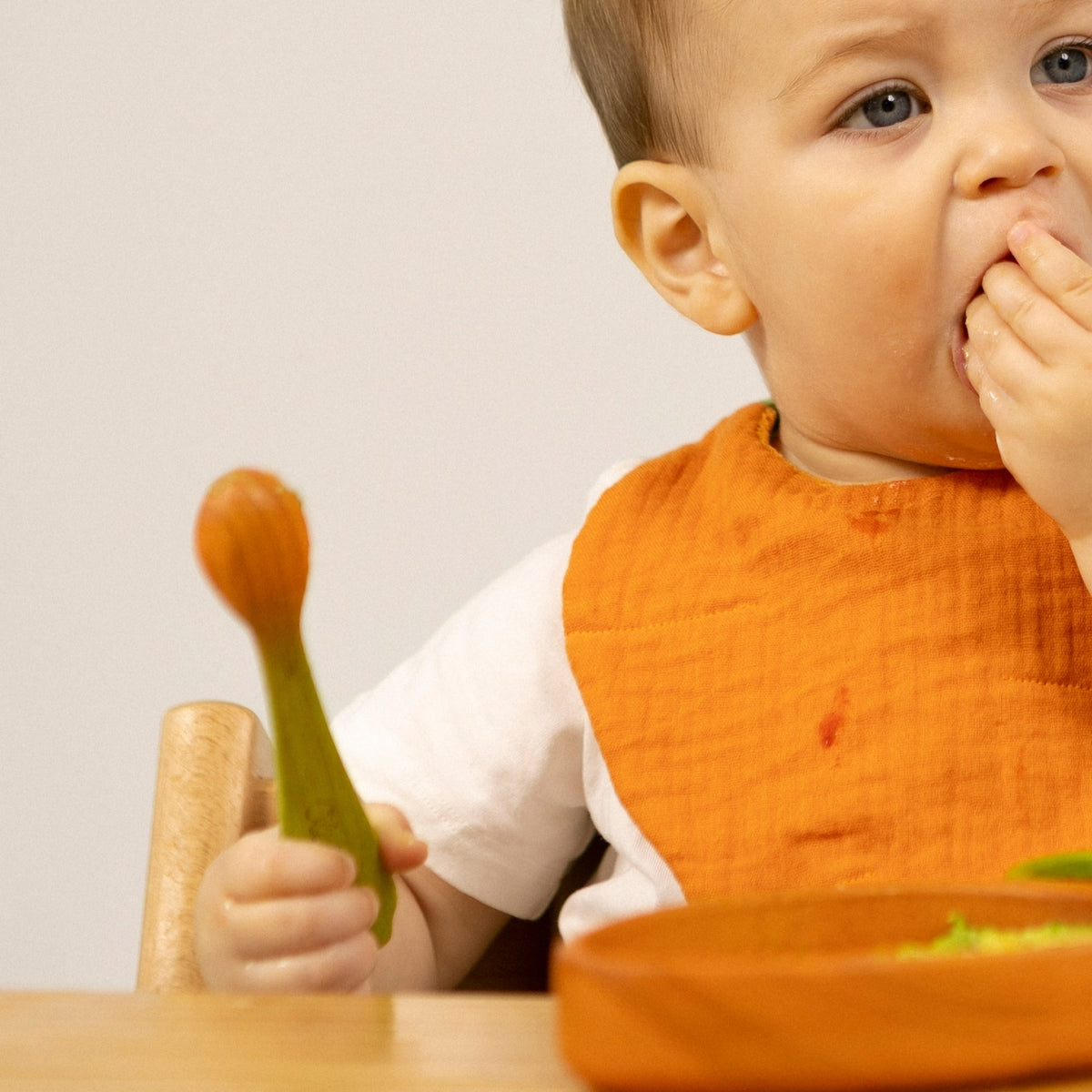 Baby in an orange bib eating with a spoon and fork at a table.