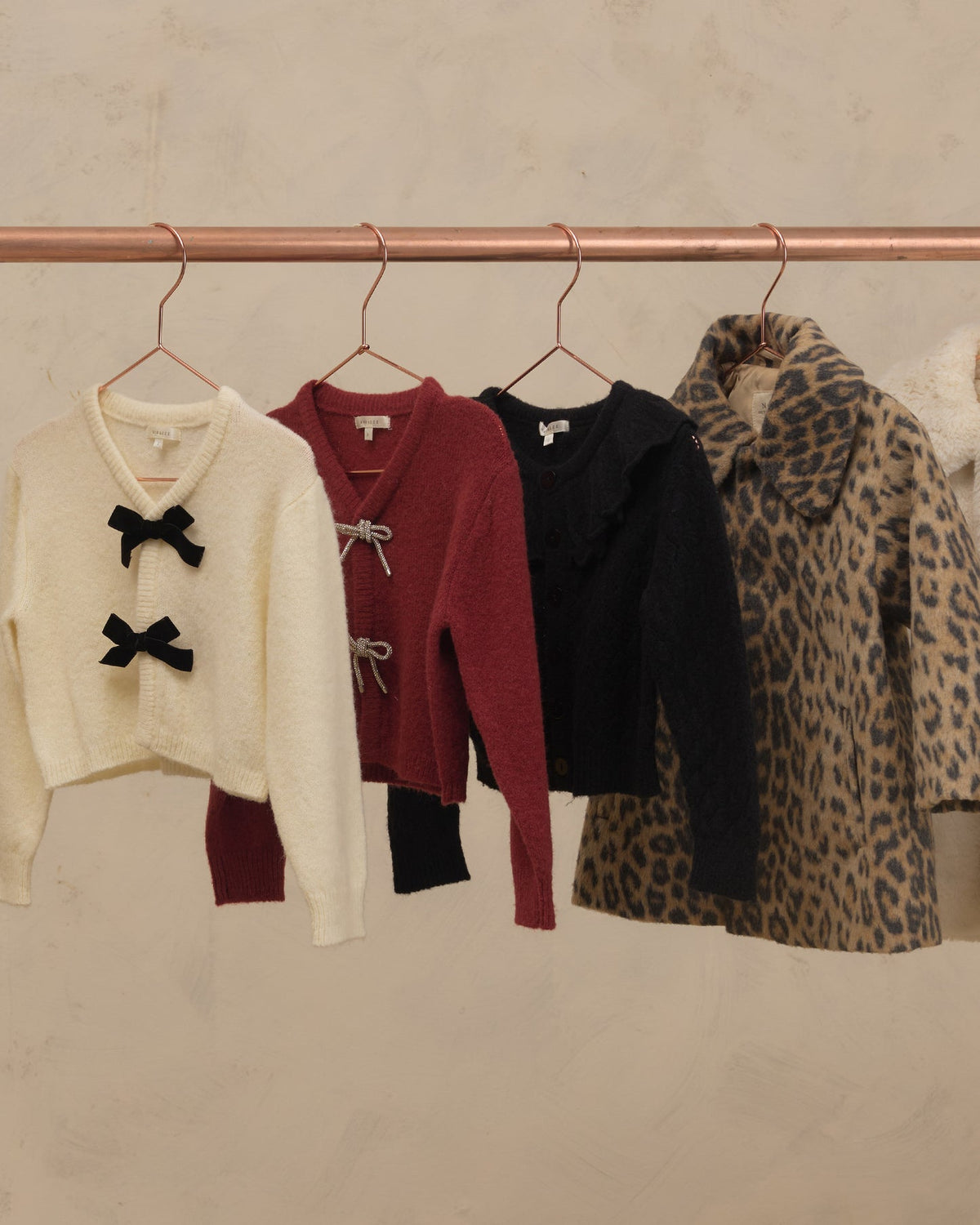Four women's knit outerwear pieces hanging on a copper-colored clothing rack against a textured beige wall. From left to right: a cream cropped sweater with black bow details, a burgundy cropped sweater with white tie details, a black ruffled cardigan, and a leopard-print coat.
