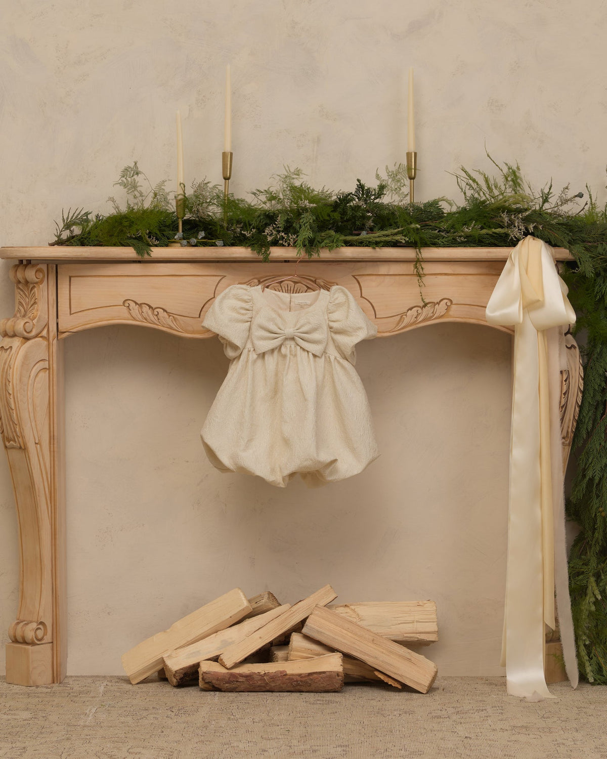 An ivory baby dress with puffed sleeves and a large bow is hanging from the center of an ornate wooden mantel. The mantel is decorated with green foliage, three tall candles, and a long cream ribbon, with stacked firewood below against a textured beige wall.