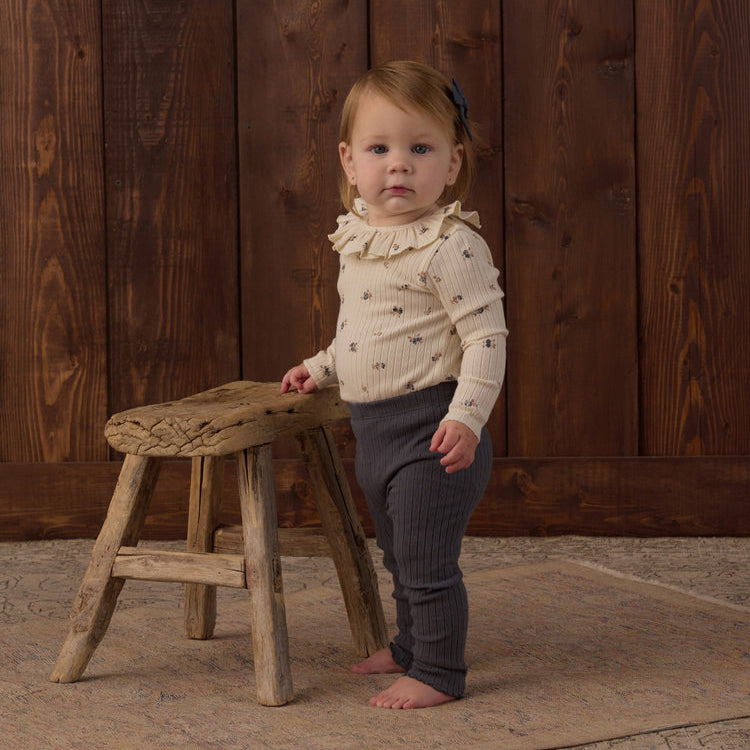 A toddler wearing Rylee + Cru Ruffle Collar Bodysuit in Natural Ditsy paired with Midnight ribbed leggings and a matching hair bow.