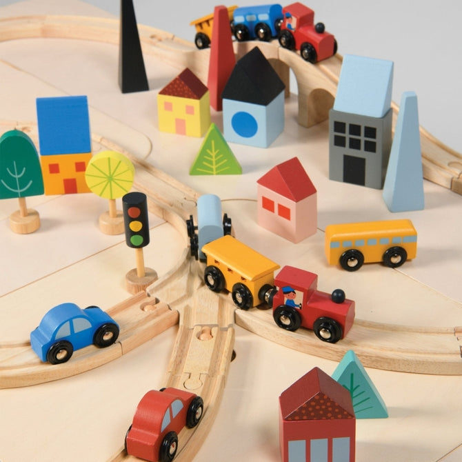 A top-down, close-up view of a wooden toy train set on a light-colored surface, featuring a curved track with a blue train engine and yellow cargo cars. Colorful wooden town buildings, including a red house, blue structures, and abstract trees, surround the tracks alongside a yellow traffic light, a red toy car, and a yellow toy truck.