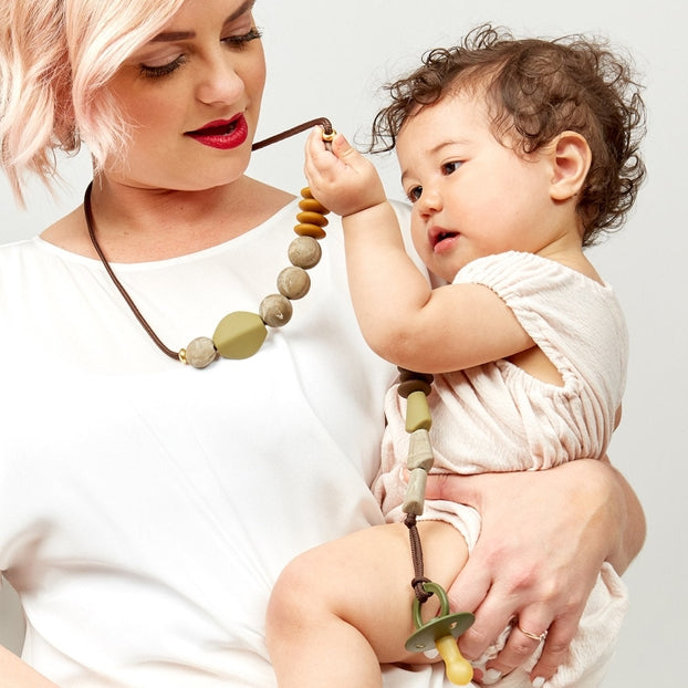 Woman holding a baby and wearing a necklace with large beads on a plain background.