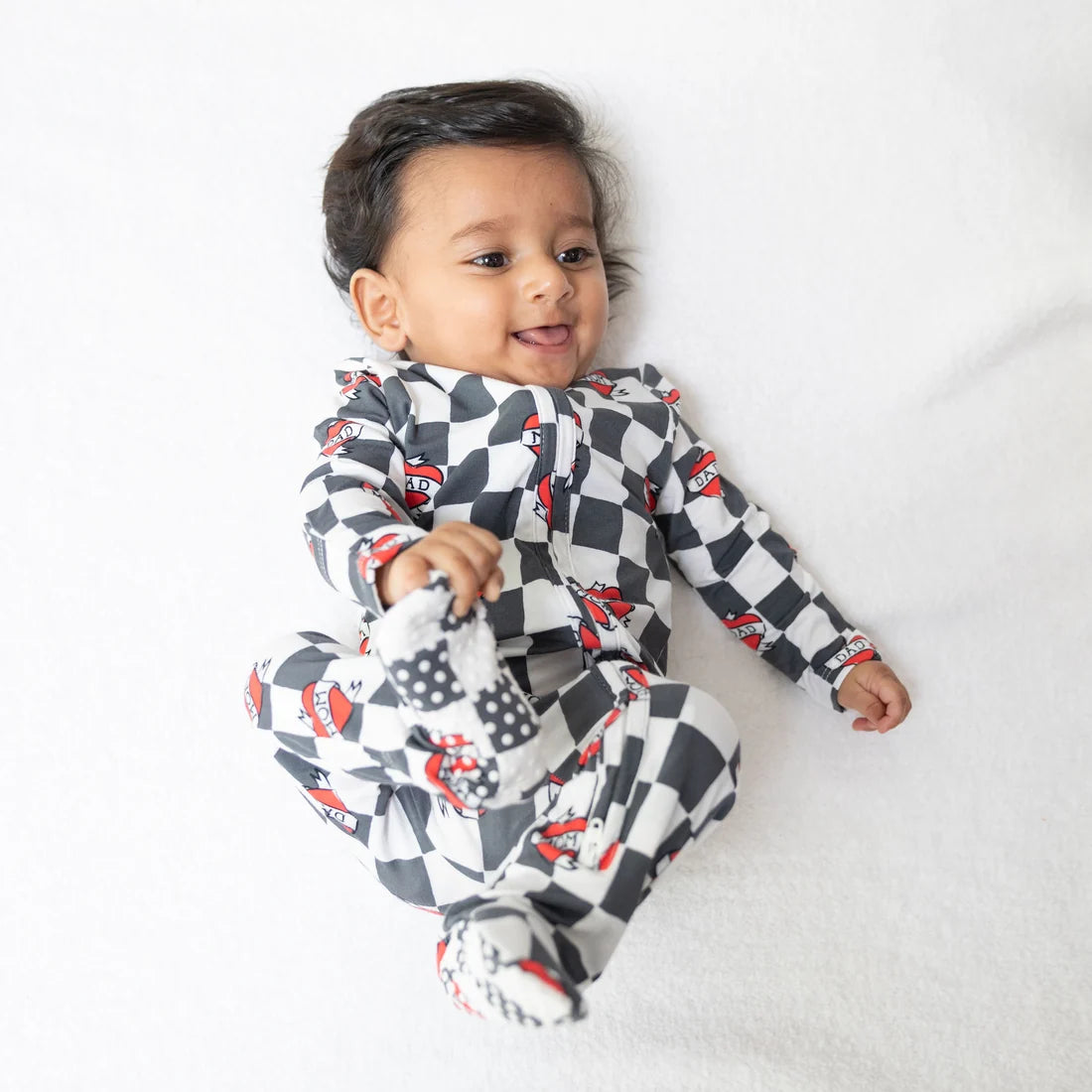 A baby lying on a white textured blanket, wearing a black and white checkered one-piece pajama with red and black cartoon car prints. The baby is holding a matching black and white checkered footed sleeper.