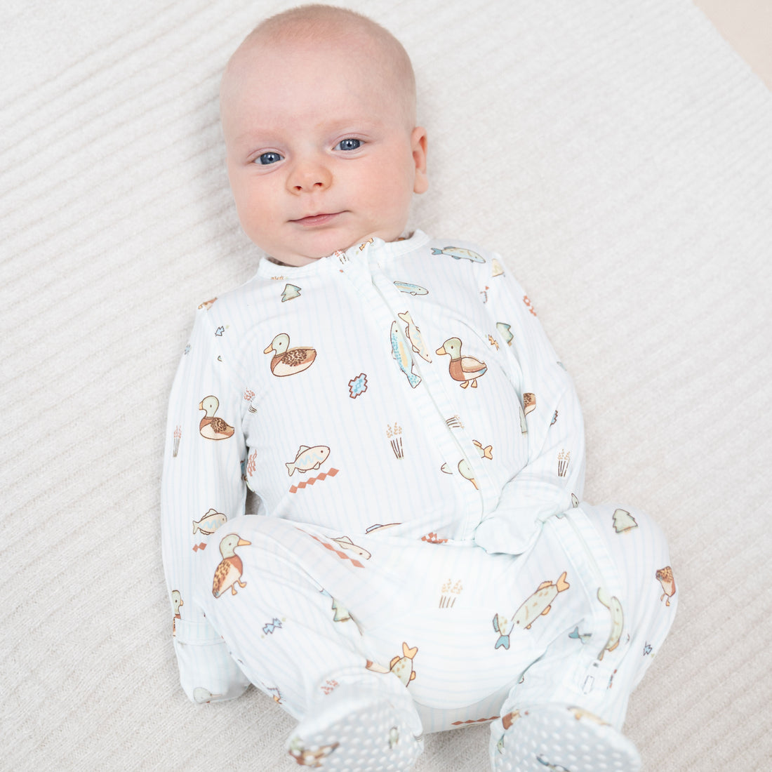 A baby lying on a textured white blanket, wearing a white footed sleep sack with a colorful duck and fish pattern.