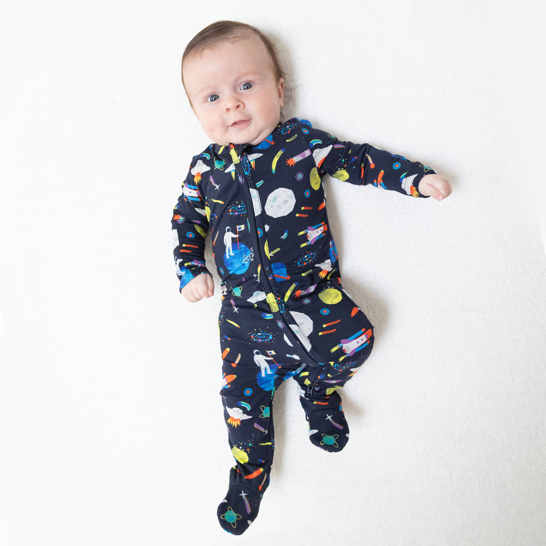 A baby lying on a white surface wearing a dark navy footed sleeper with a colorful space-themed print of planets, rockets, and stars.