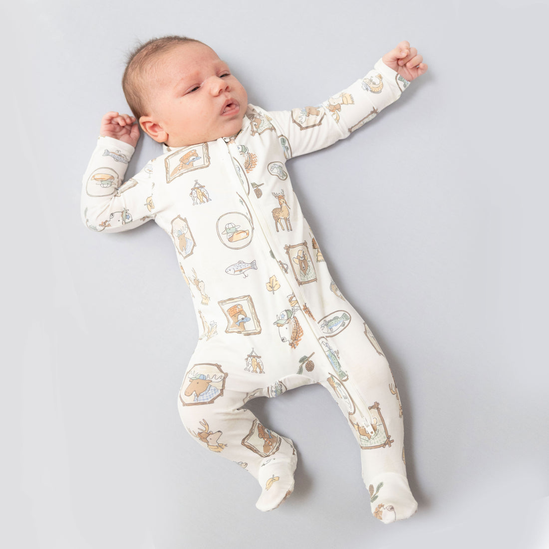 A sleeping newborn baby wearing a long-sleeve footed sleeper with a whimsical animal and house pattern, lying on a light gray background.