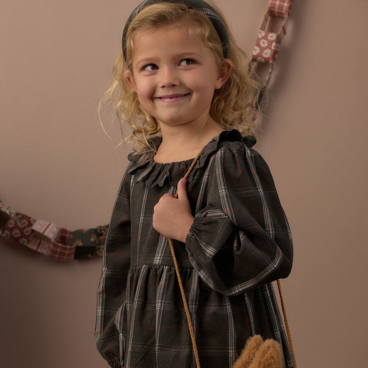A young girl with blonde curly hair wearing a dark gray plaid long-sleeve dress and a dark headband, holding a brown star-shaped purse against a warm beige background.
