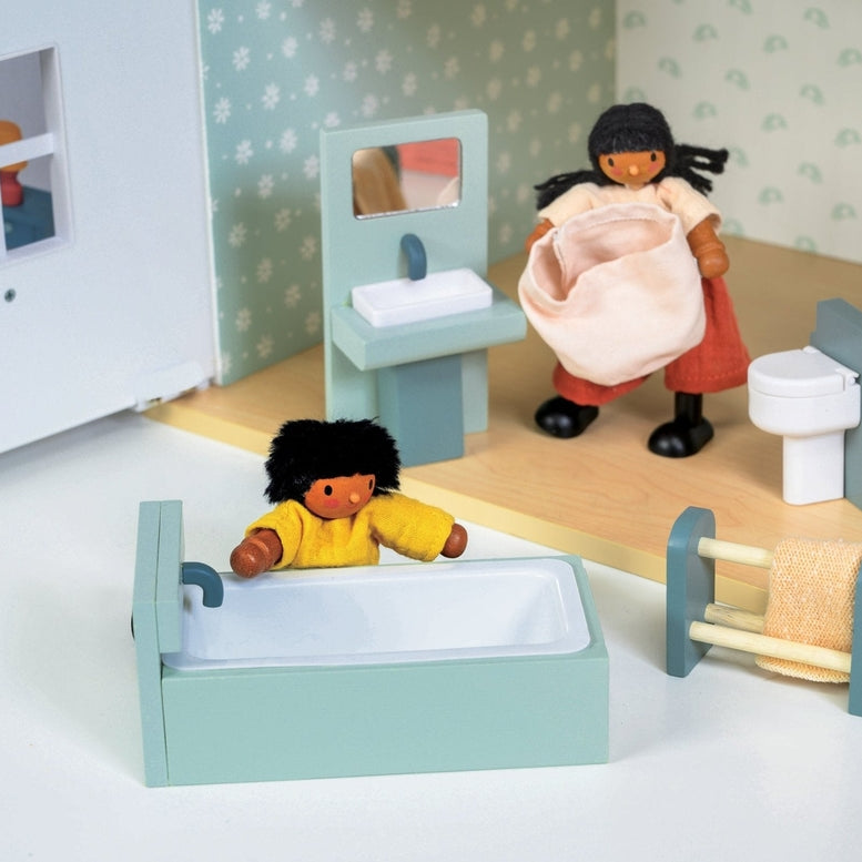 A lifestyle product photo of the Wooden Bathroom Furniture set by Mentari set up in a miniature dollhouse bathroom. One doll with a yellow shirt relaxes in the mint-green and white bathtub, while another doll standing near the sink unit carries a small pink fabric laundry basket.