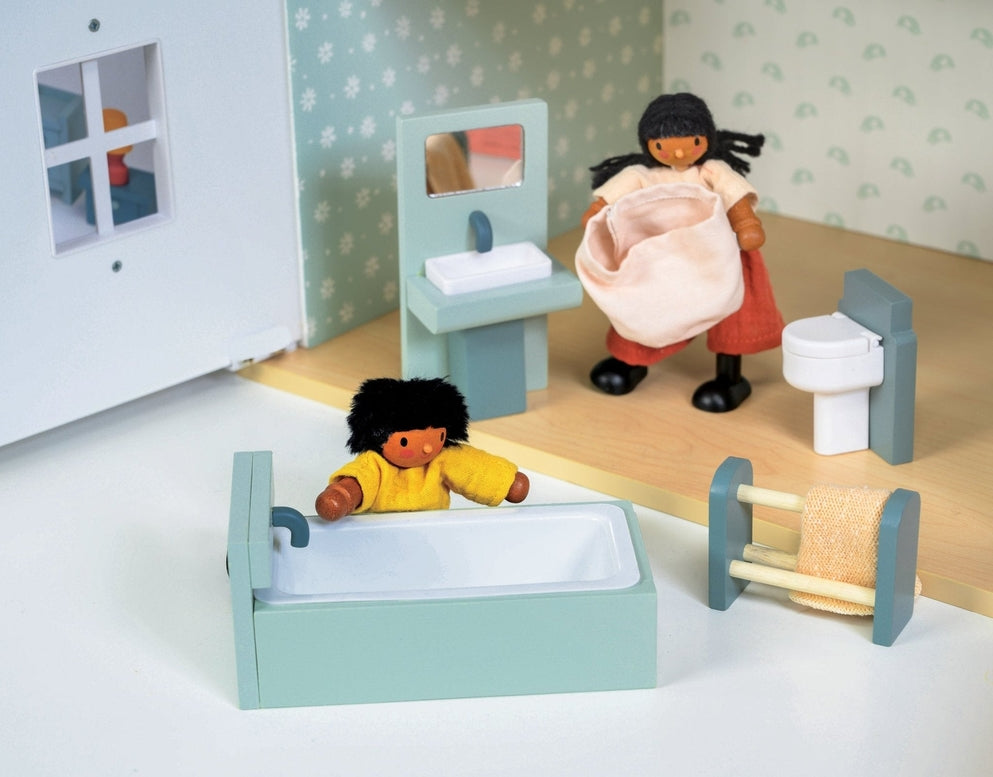 A lifestyle product photo of the Wooden Bathroom Furniture set by Mentari set up in a miniature dollhouse bathroom. One doll with a yellow shirt relaxes in the mint-green and white bathtub, while another doll standing near the sink unit carries a small pink fabric laundry basket.